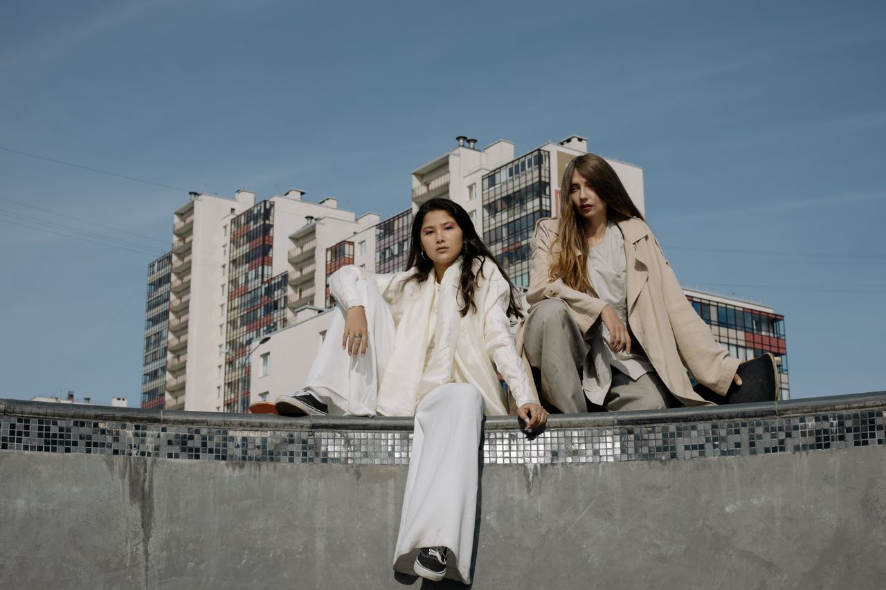 Home Two fashionable women sitting outdoors against a backdrop of urban buildings.